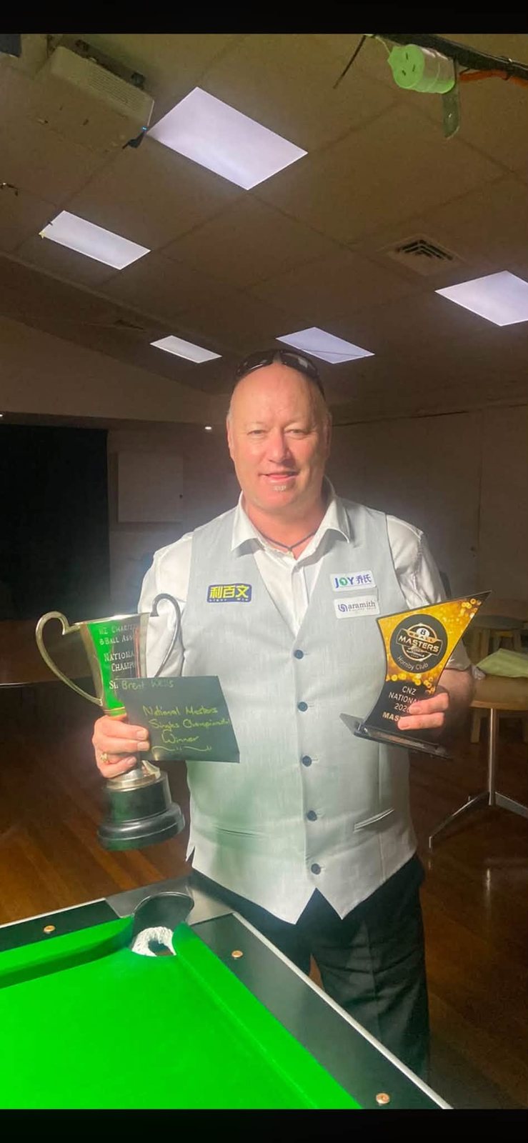 Pool player holding two trophies beside a pool table after a national masters singles final